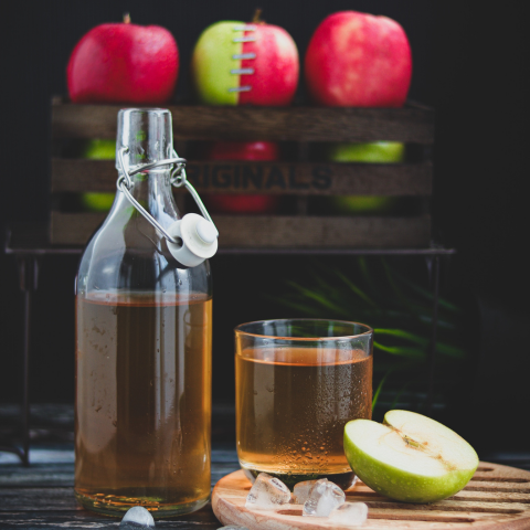 bottle of apple cider vinegar open with a glass to the right and a green apple cut in half. int he background, you can see red and green apples stacked with half a red and half a green apple stapled together to show both kinds in the vinegar